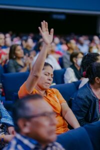 Woman in audience raising her hand during a public gathering, showing engagement.